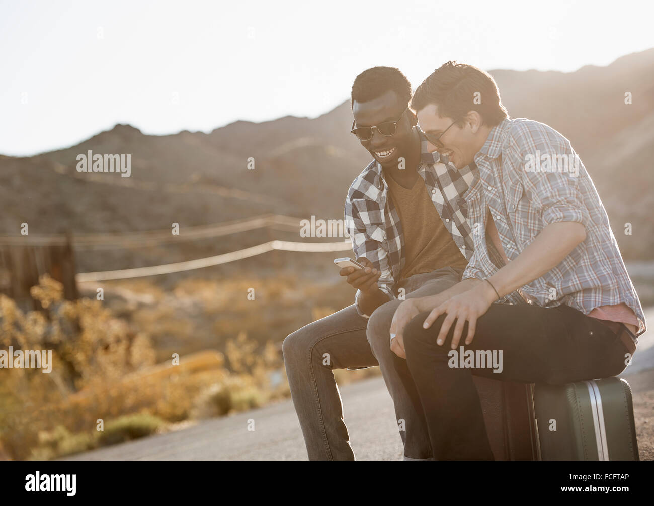 Due giovani uomini seduti sul ciglio della strada sulla loro casi utilizzando un telefono intelligente. Foto Stock