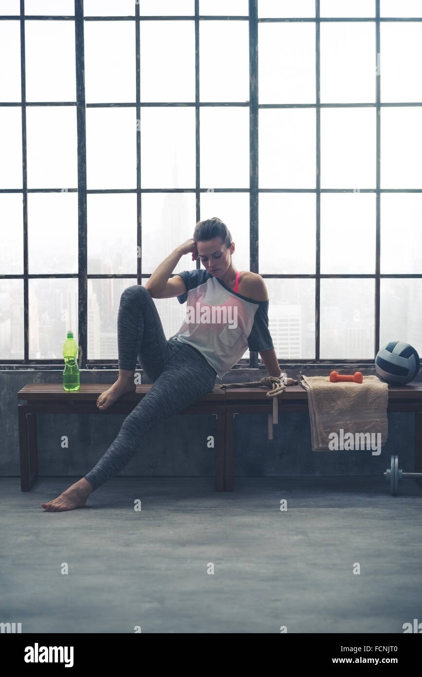 Un accoppiamento, donna sana in allenamento marcia è seduta su una panchina, appoggiando un gomito sul suo ginocchio piegato e la sua testa sulla sua mano. Lei è Foto Stock