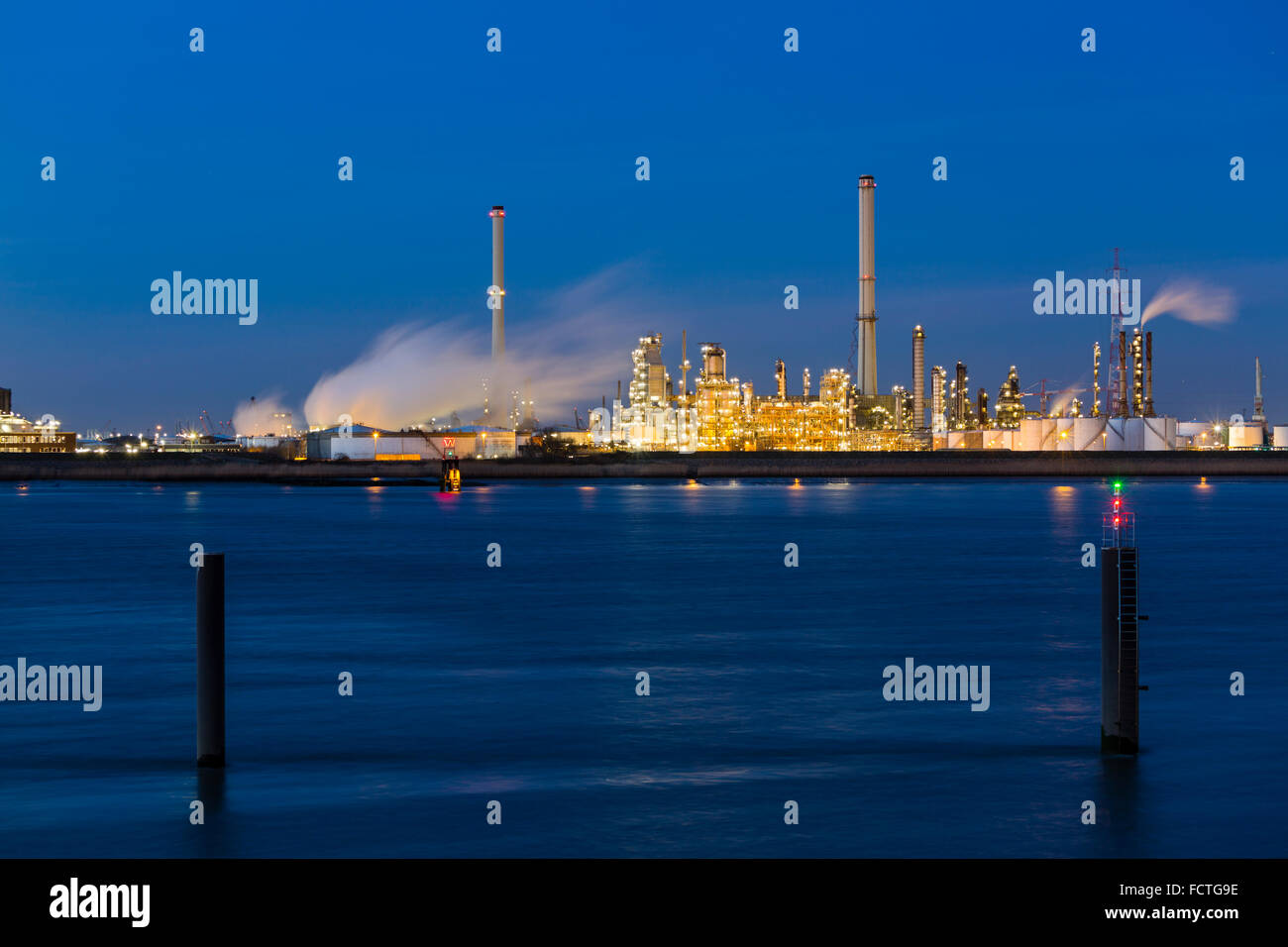 Vista sul fiume Schelda a una grande raffineria di petrolio con night blue sky, illuminazione e di vapore, due poli in primo piano. Foto Stock