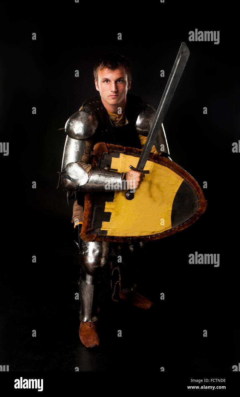 Studio shot del giovane uomo vestito da cavaliere medievale armato di spada e scudo in pronto a combattere la posizione su sfondo nero Foto Stock