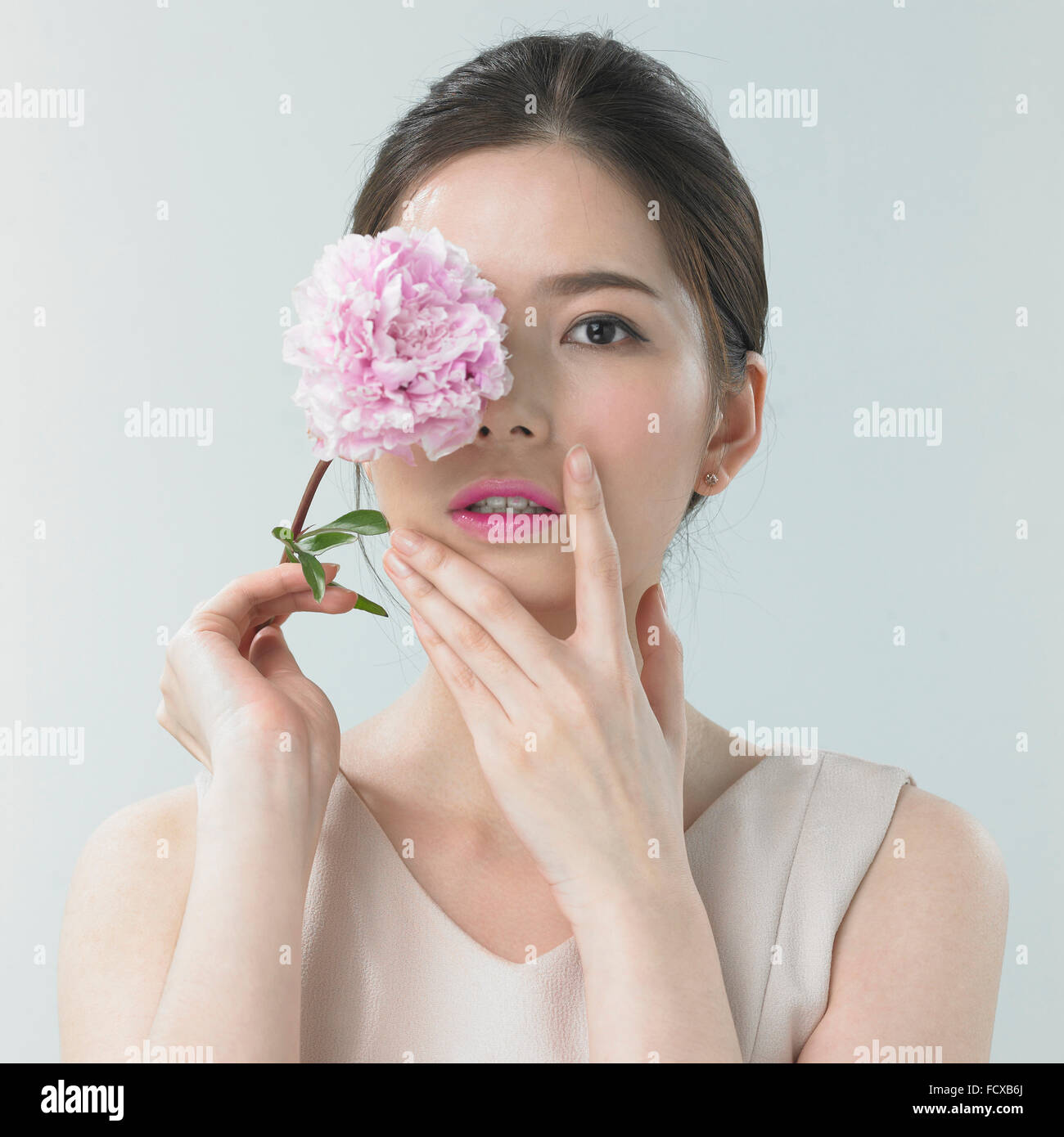 Donna con labbro di rosa per il make-up tenendo un fiore rosa che copre il suo occhio con l'altra mano sul suo viso Foto Stock