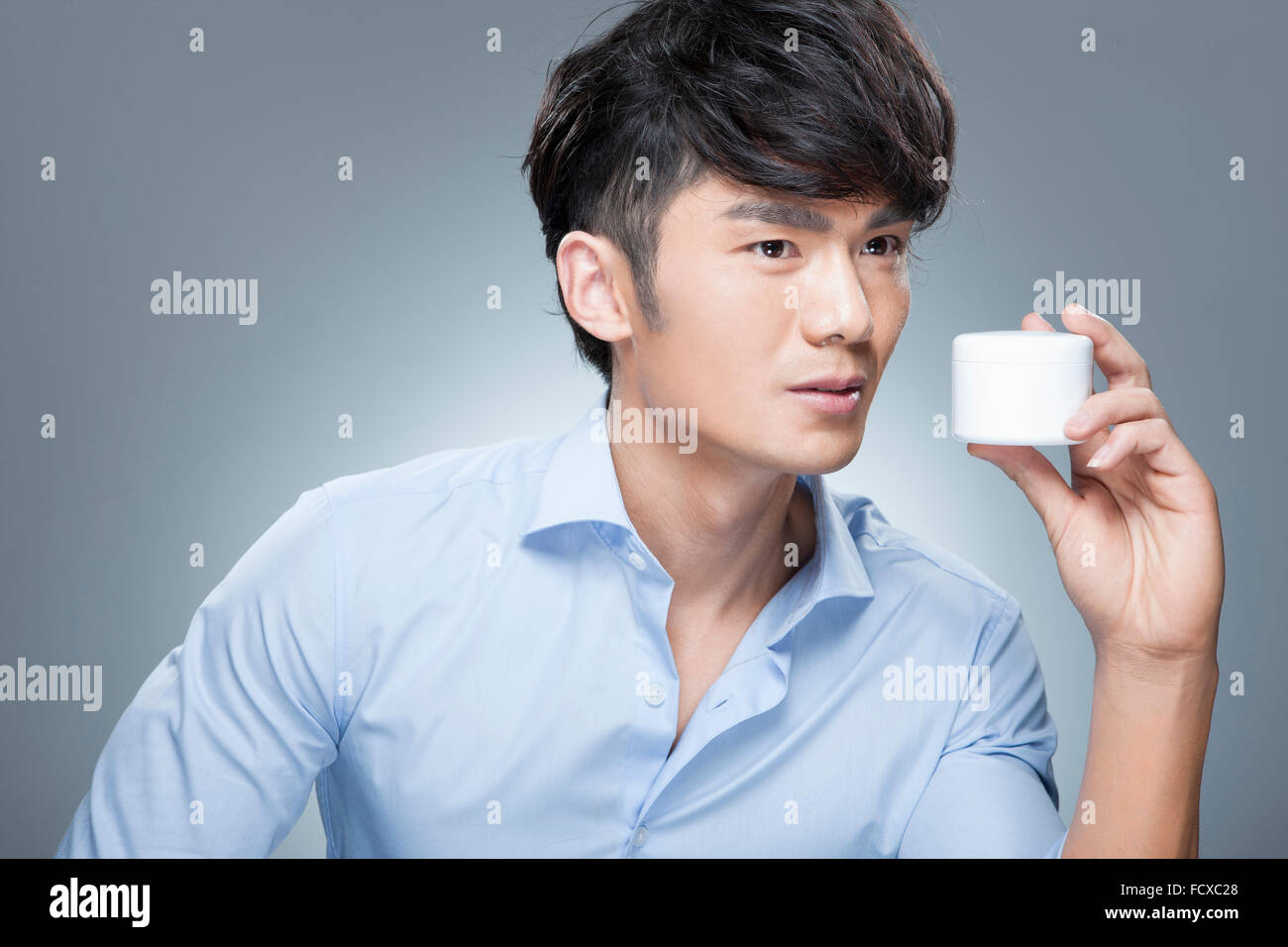 Uomo in camicia blu tenendo una crema per uomo di prodotto per la cura della pelle accanto al suo viso e lo sguardo rivolto Foto Stock