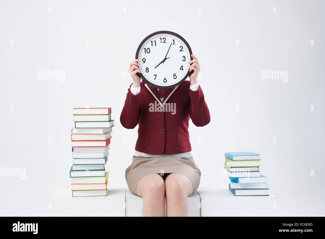 High School girl in uniformi di scuola tenendo un orologio che copre il volto e la seduta tra pile di libri Foto Stock