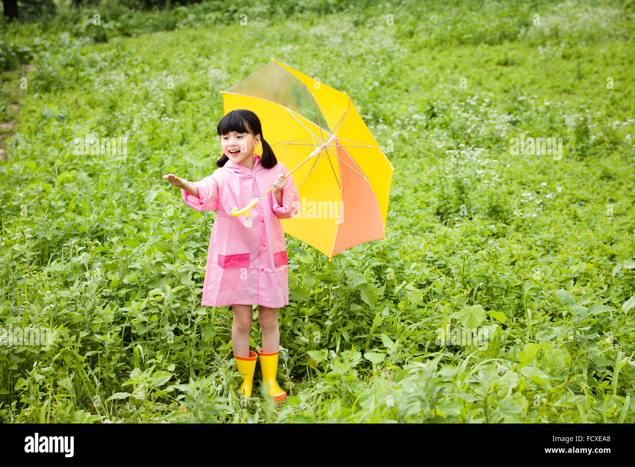 Giovane ragazza in cappotto di pioggia e pioggia stivali sotto Ombrello giallo il suo stiro in avanti il braccio nel campo Foto Stock