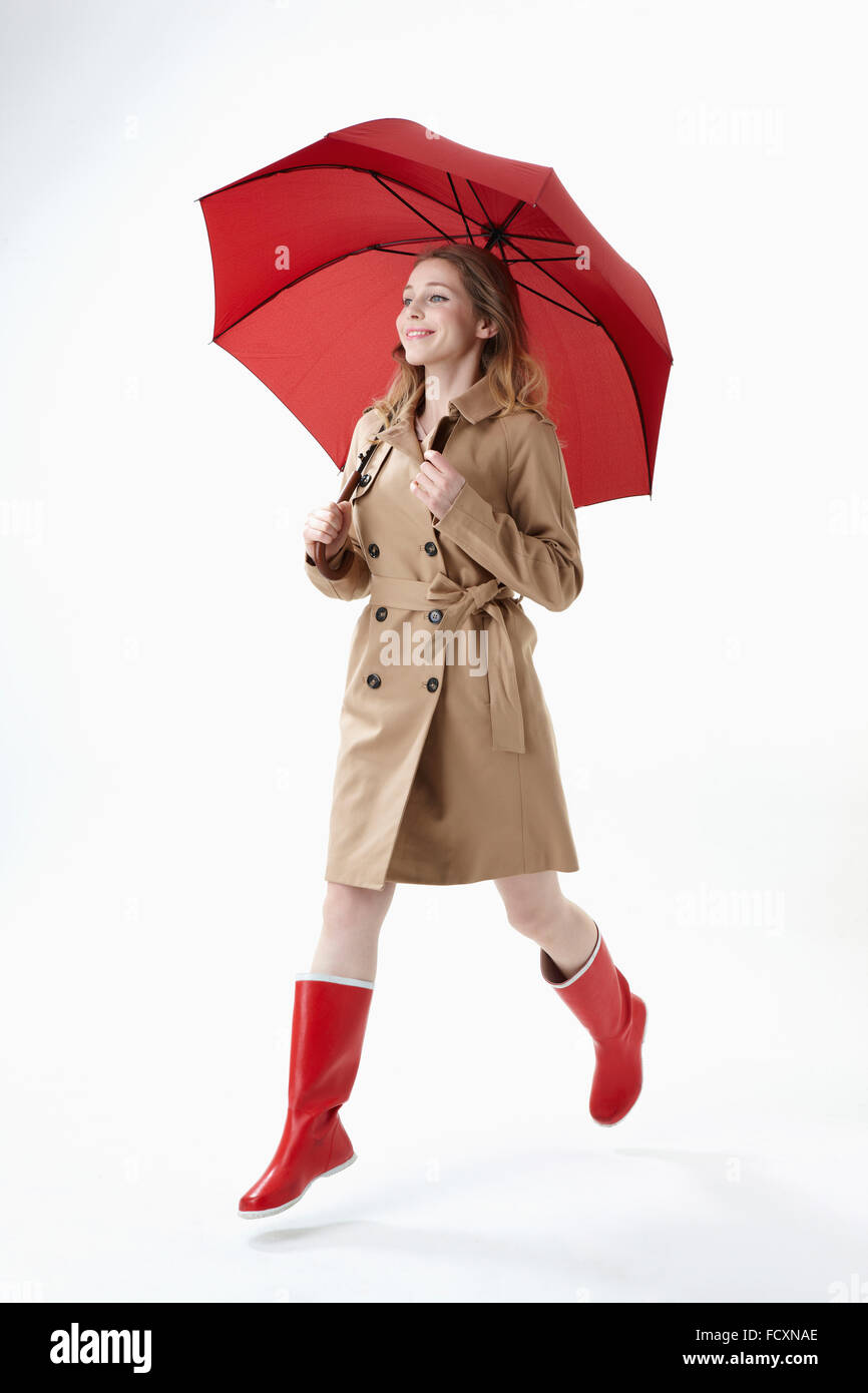 Giovane donna sorridente indossando Ombrellone rosso, trench e stivali rossi in esecuzione Foto Stock
