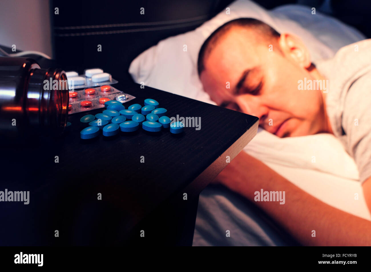 Primo piano di alcune diverse pillole nel comodino e un giovane uomo che stabilisce a faccia in giù nel letto con gli occhi chiusi, di notte Foto Stock