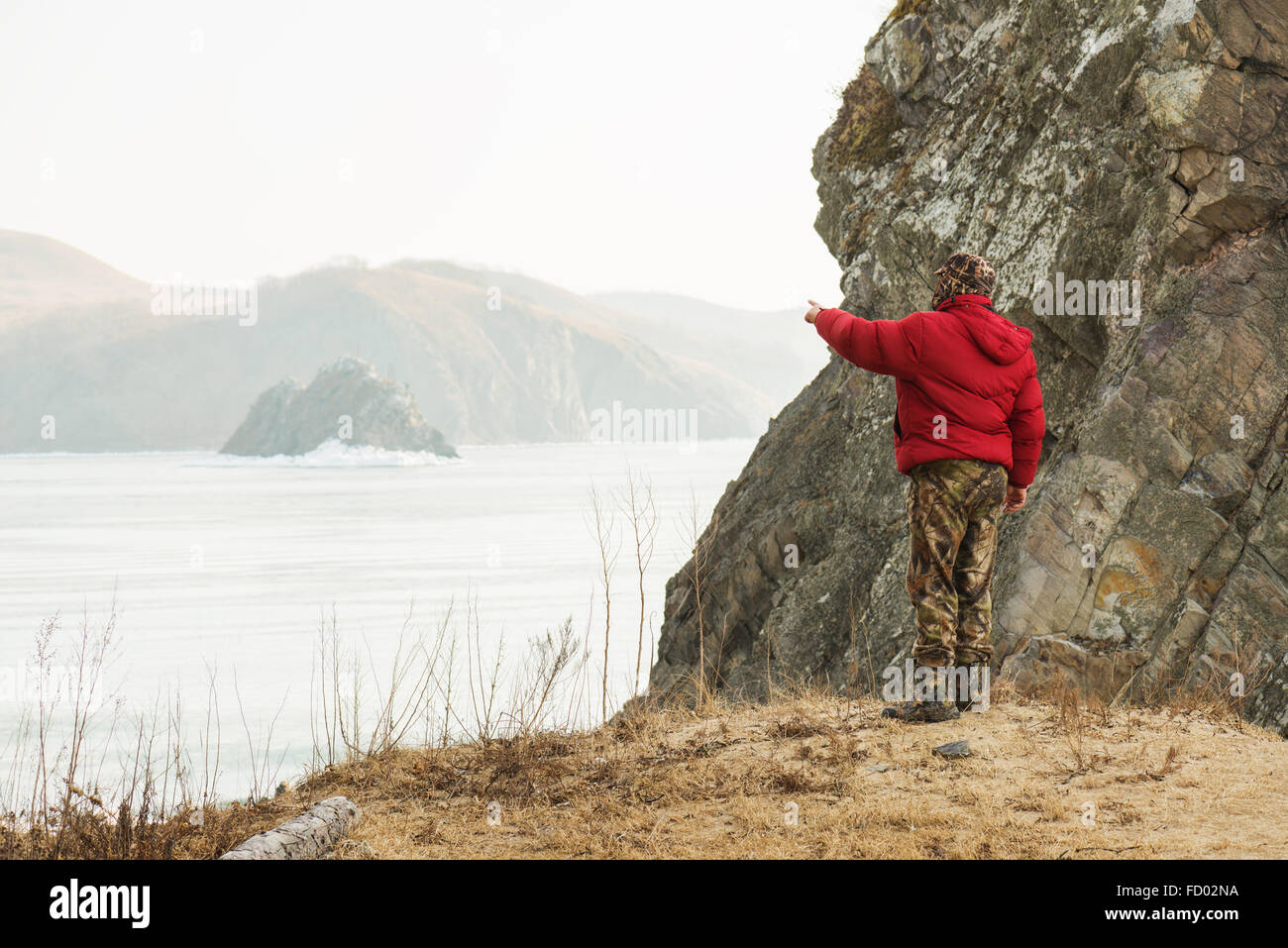 Uomo in tuta mimetica e giacca rossa permanente sulla spiaggia o sul lago e indica a mano in direzione di caccia o di movimento. Foto Stock