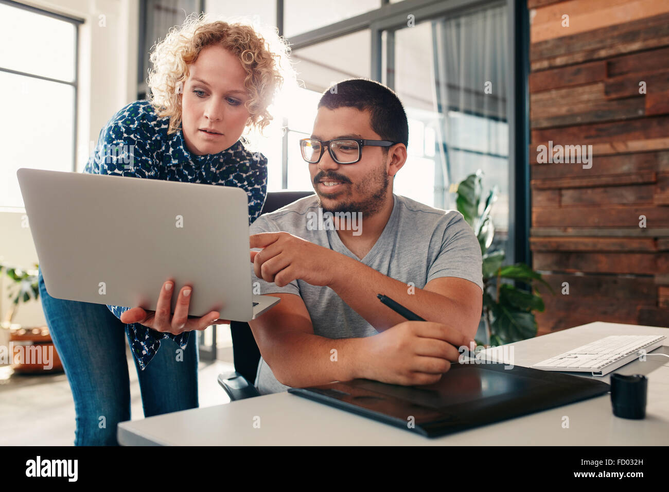 Ritratto di giovane uomo seduto alla sua scrivania puntando al portatile nella mani di un collega femmina, maschio graphic designer aiutano a femmina Foto Stock