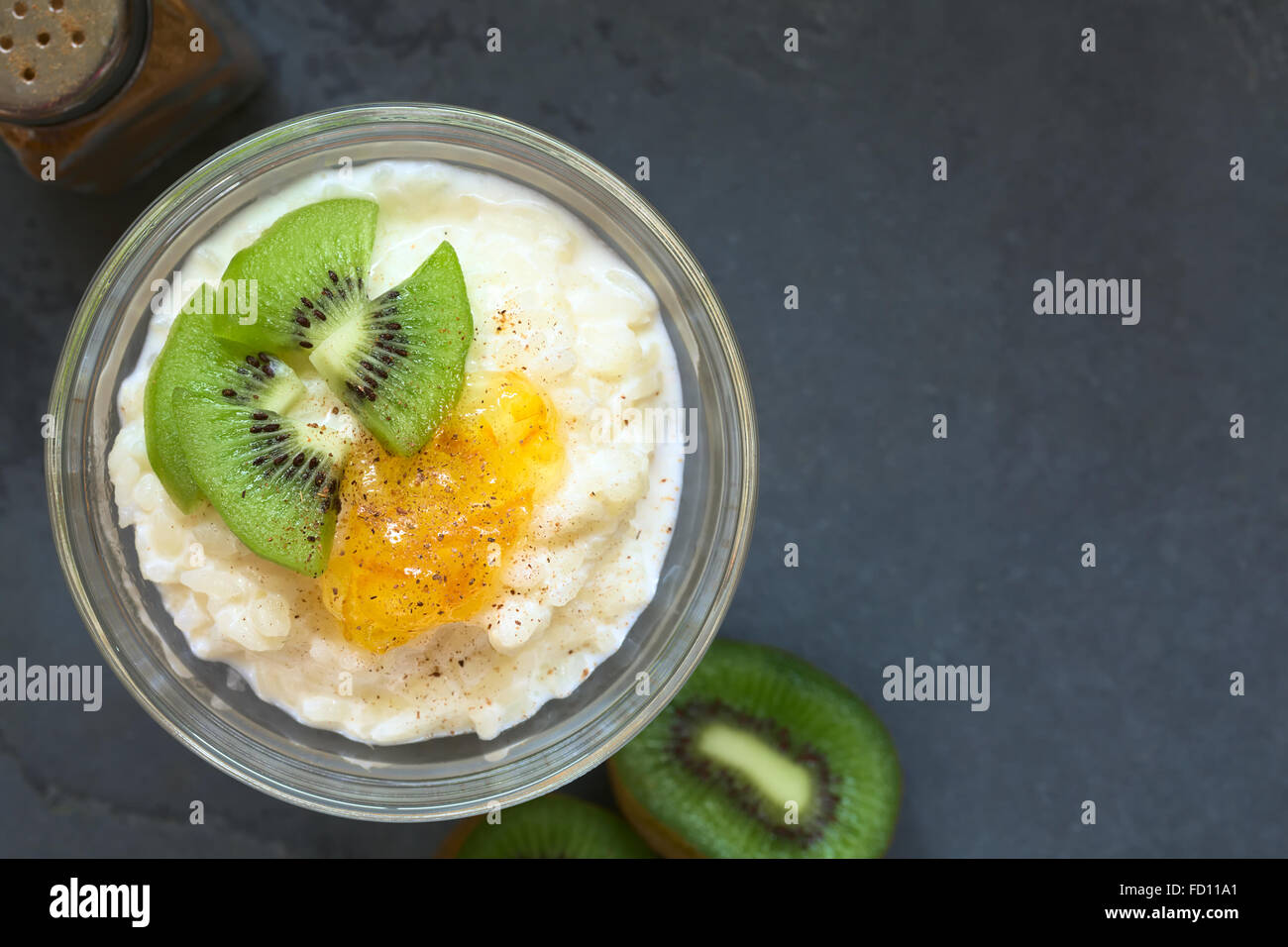 Budino di riso con pezzi di kiwi, marmellata di arancio e cannella in piccola ciotola di vetro, fotografato overhead su ardesia con luce naturale Foto Stock