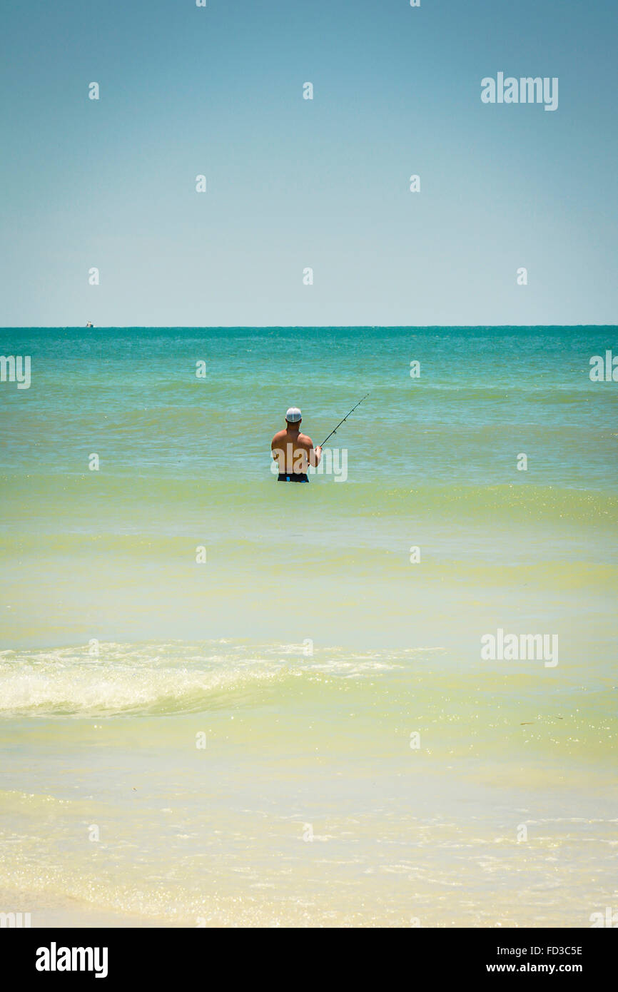 Una vista posteriore di un uomo solitario surf pesca con un asta di luce in waist deep acque blu turchese contemplare il bellissimo paesaggio marino Foto Stock