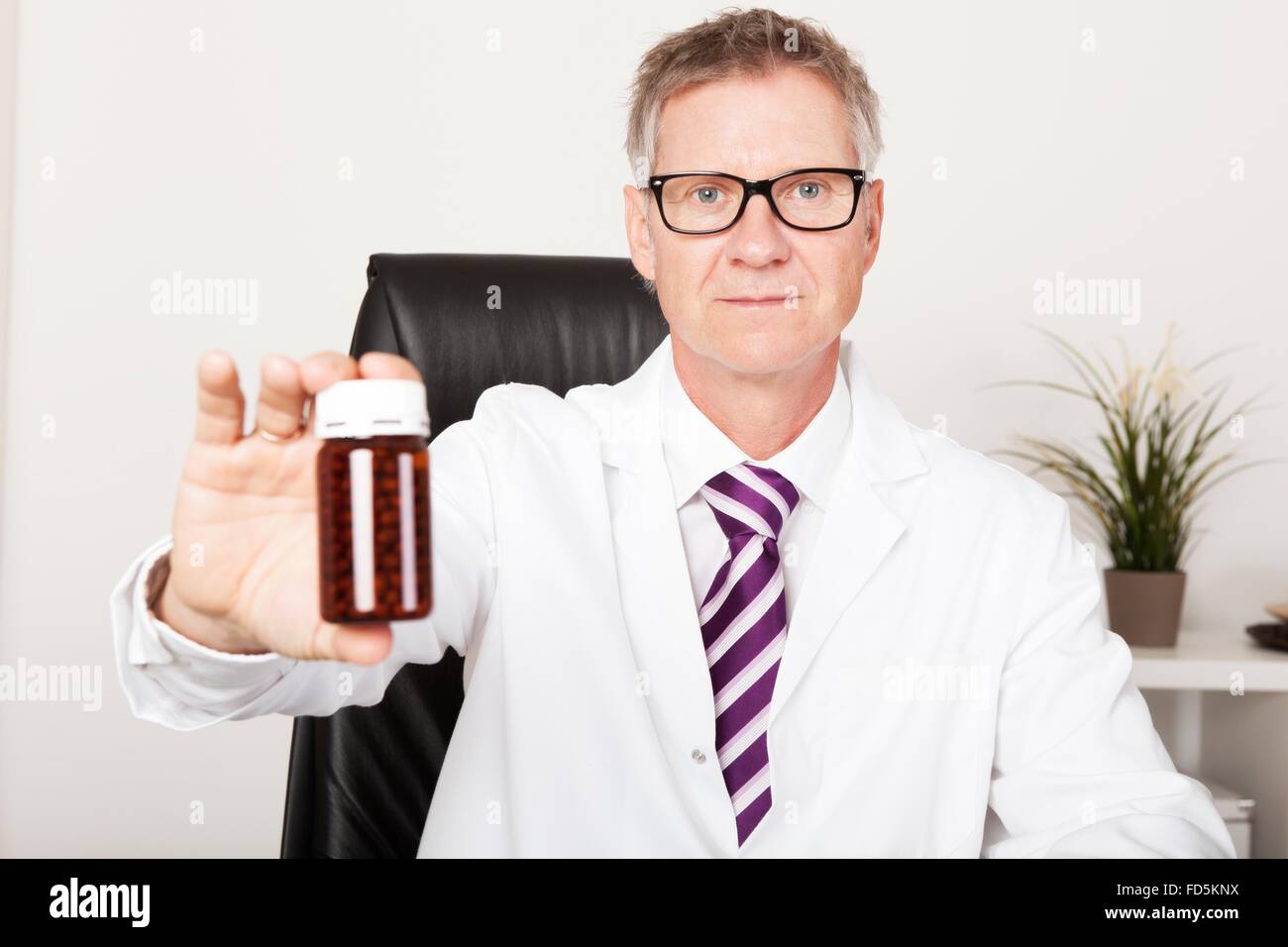 Metà maschio invecchiato o un farmacista o medico tenendo fuori un marrone non marcato la bottiglia di plastica di pillole come si siede a una scrivania in un ufficio Foto Stock