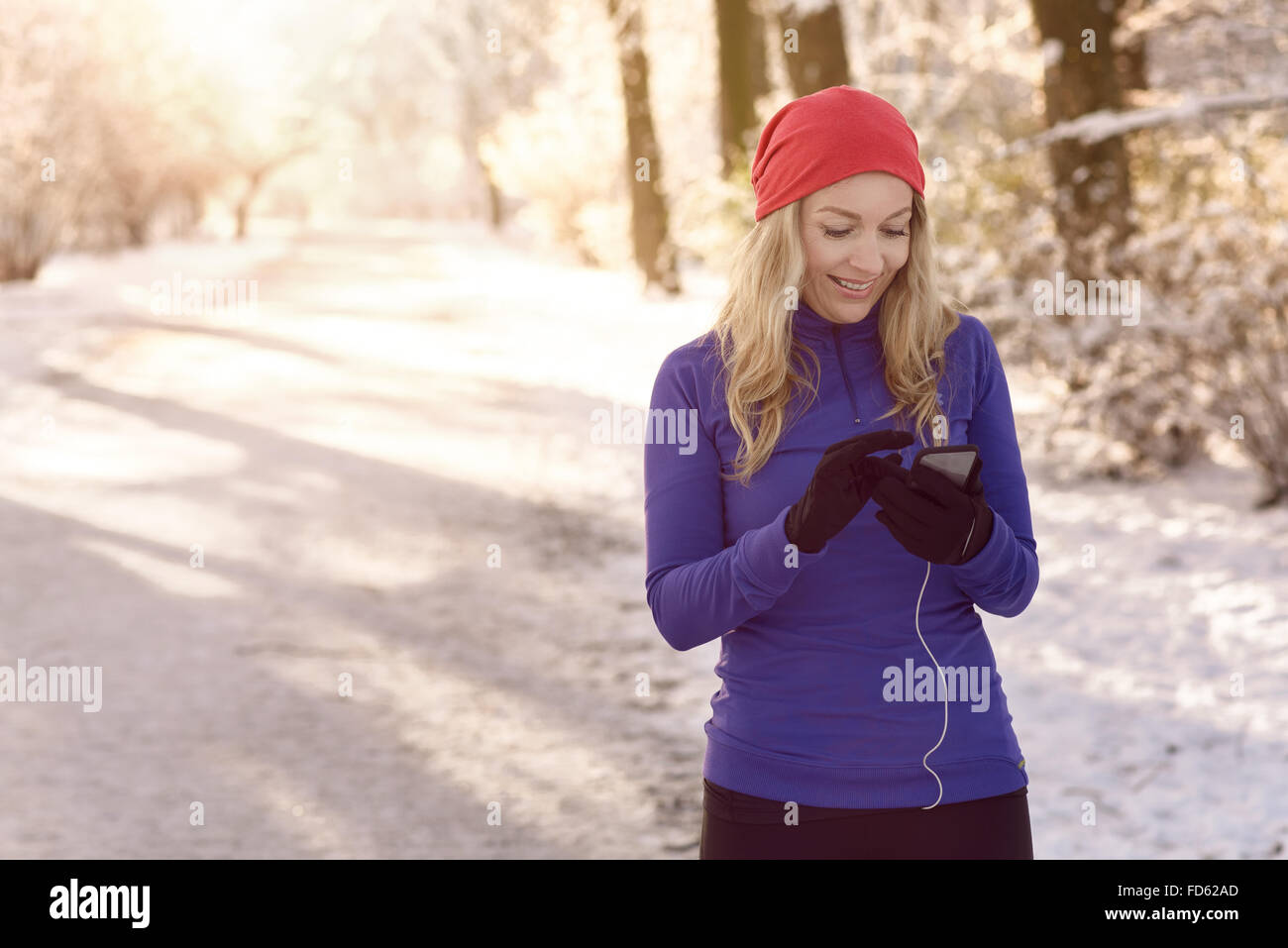 Donna controllo lei pone mobile per i messaggi di testo con un sorriso come lei gode di una salutare passeggiata invernale attraverso un parco innevato in Foto Stock