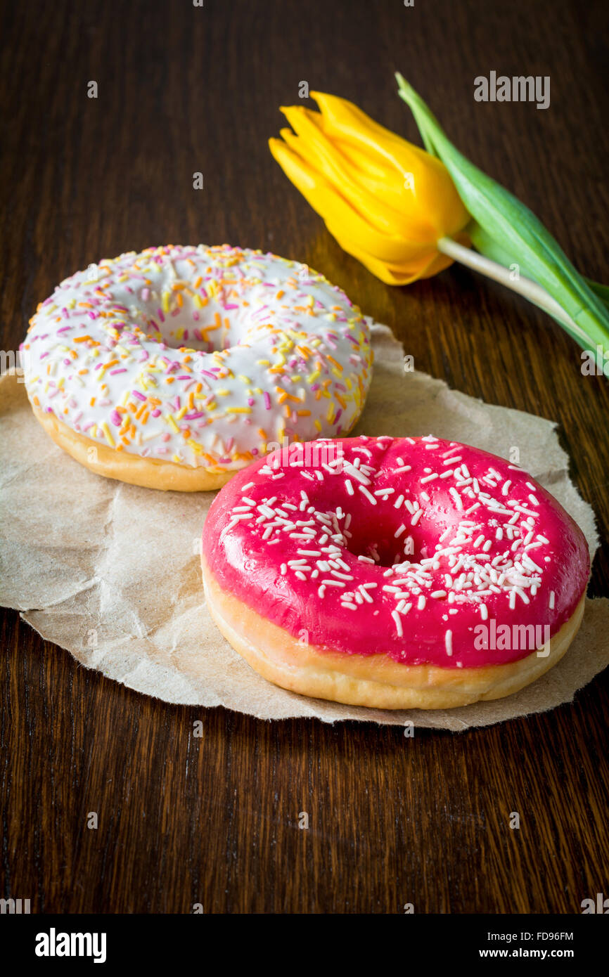 Due ciambelle smaltate e tulipano giallo fiore marrone sul tavolo di legno. La prima colazione dolce Foto Stock