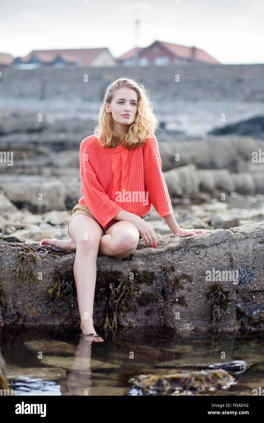 Bella giovane donna seduta su una roccia sulla spiaggia. Lei è a piedi nudi e indossa un abbigliamento casual. Foto Stock