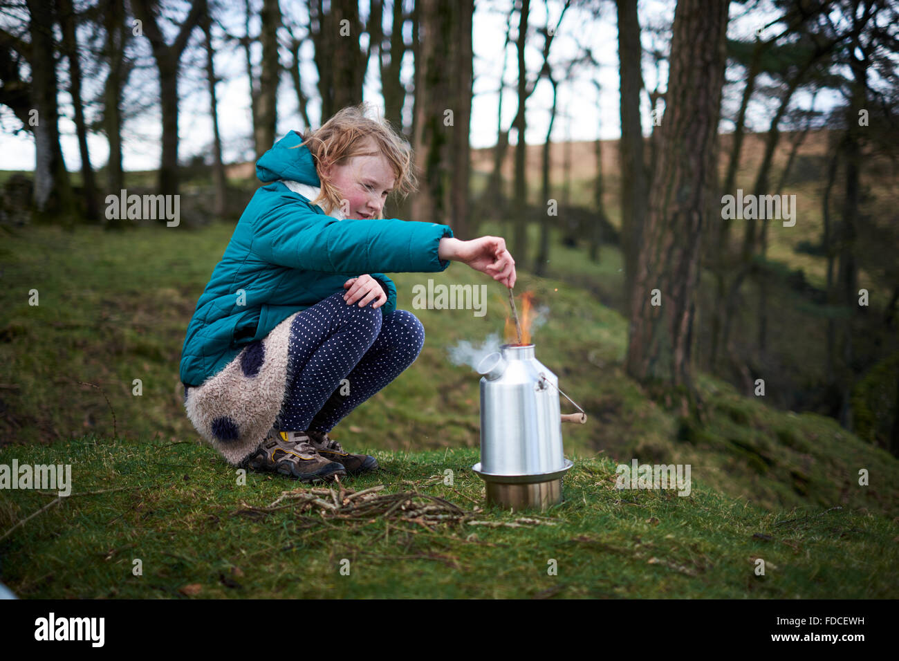 Una ragazza utilizzando un bollitore Kelly Stufa per cucinare all'aperto sorridente e aggiunta di legna per la stufa. Foto Stock