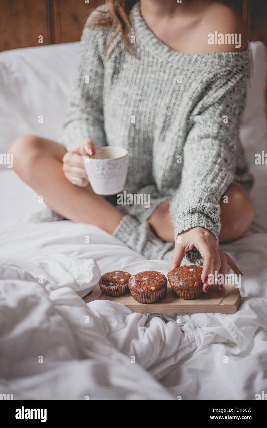 Donna seduta sul letto con la tazza di tè e caffè e muffin Foto Stock