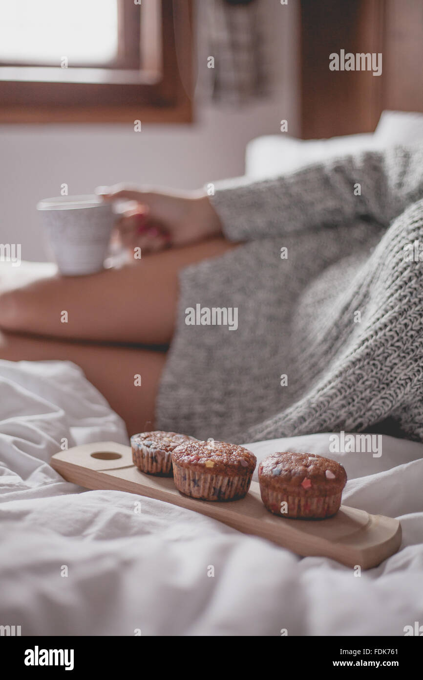Donna seduta sul letto con la tazza di tè e caffè e muffin Foto Stock