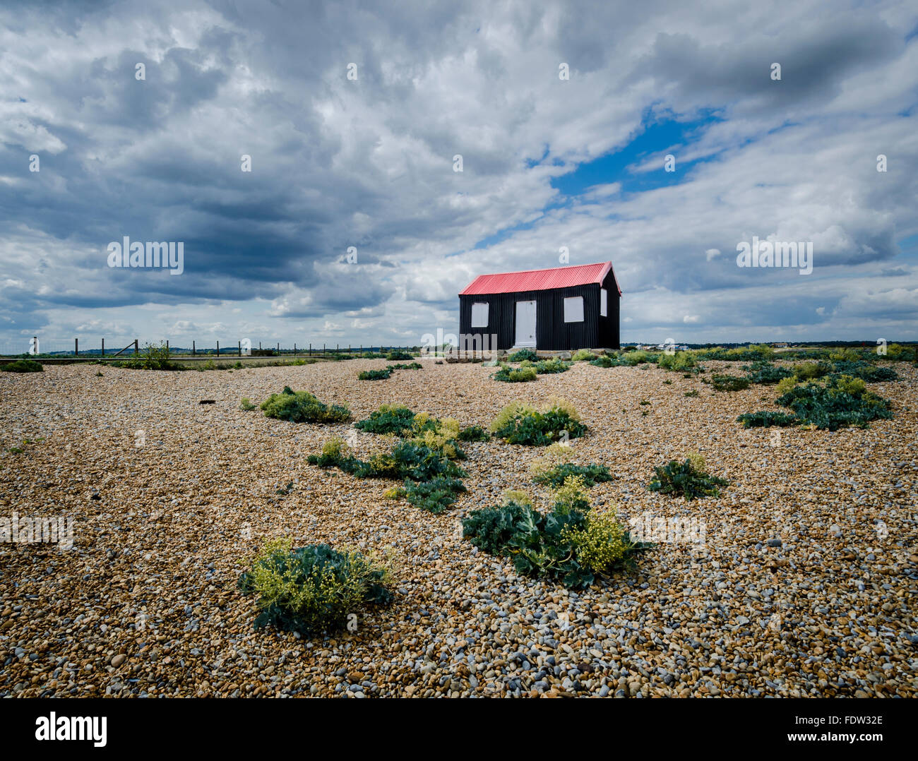 Vista di un nero capannone ondulato con un tetto rosso e ciottoli in primo piano, a Rye Harbour, East Sussex, Regno Unito Foto Stock