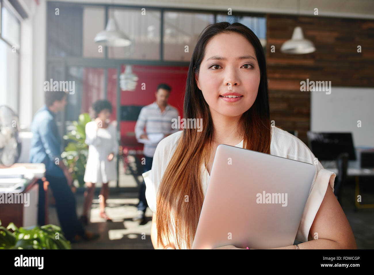Ritratto di giovane professionista creativo holding portatile in ufficio. Giovane donna asiatica durante il lavoro con i colleghi per discutere in backgr Foto Stock