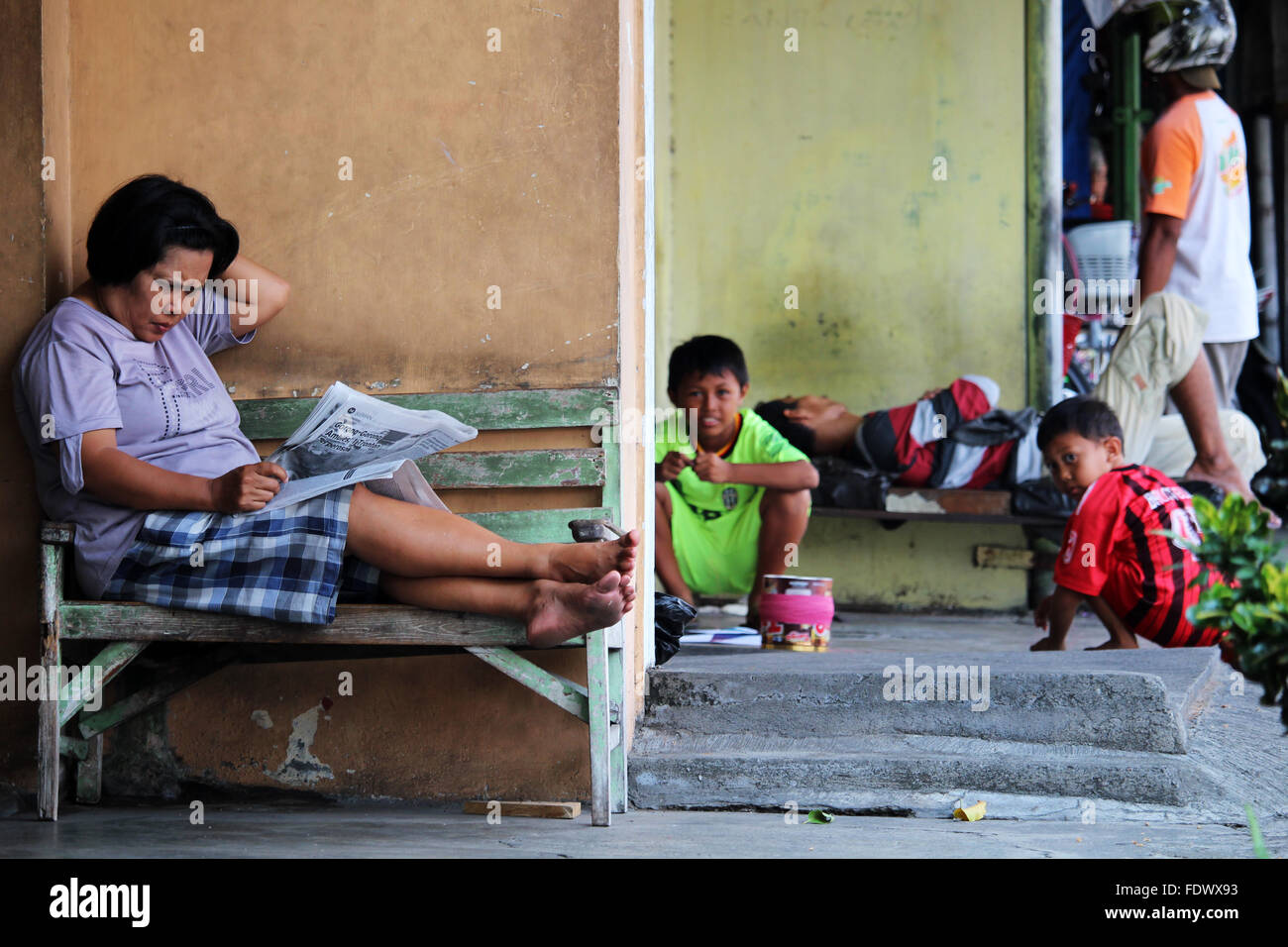 Un senior lady leggendo un giornale e, dietro, bambini che giocano. Una normale scena di vita nel centro di Yogyakarta, Indonesia Foto Stock