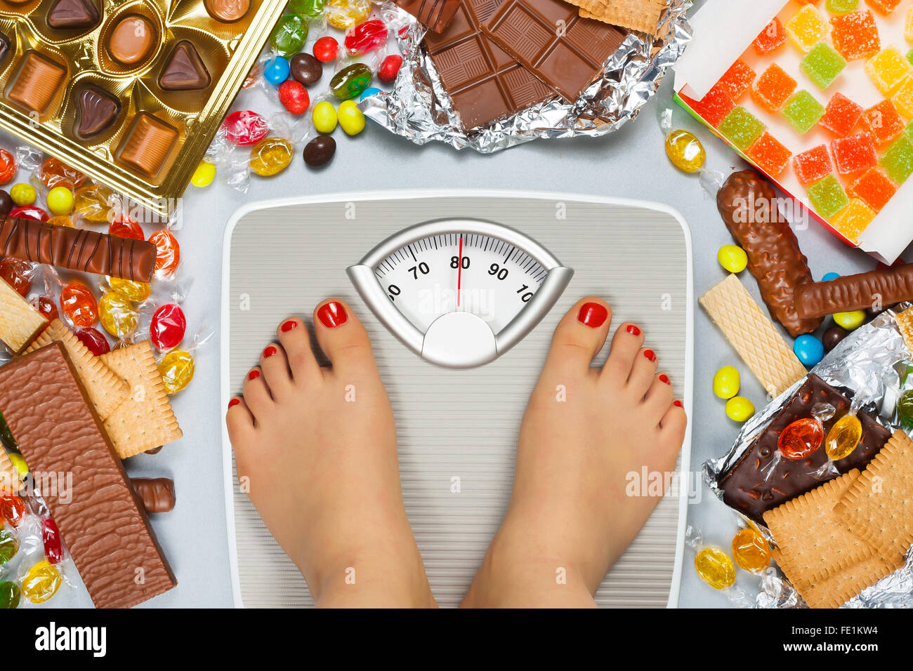 Dieta malsano - il sovrappeso. Piedi su una bilancia da bagno e il cioccolato e la gelatina a cubetti, caramelle, barrette di cioccolato, cookie, ciambelle Foto Stock