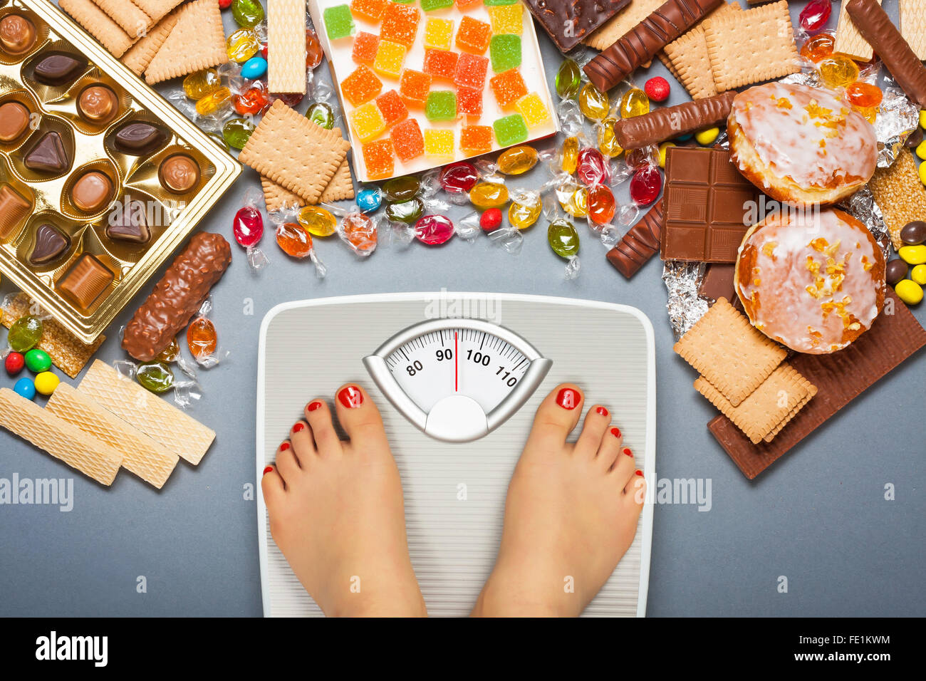 Dieta malsano - il sovrappeso. Piedi su una bilancia da bagno e il cioccolato e la gelatina a cubetti, caramelle, barrette di cioccolato, cookie, ciambelle Foto Stock