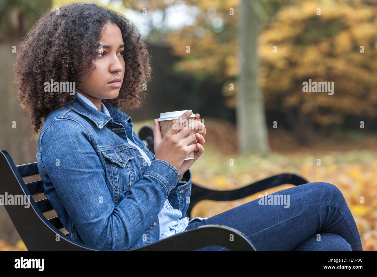Bellissima gara di misto americano africano ragazza adolescente femmina giovane donna a bere il caffè da asporto al di fuori seduta su una panchina nel parco Foto Stock