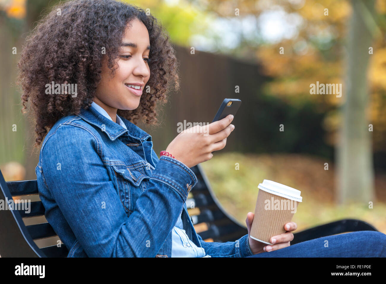 Bella felice razza mista americano africano ragazza adolescente femmine giovane donna sorridente a bere il caffè da asporto telefono cellulare SMS Foto Stock