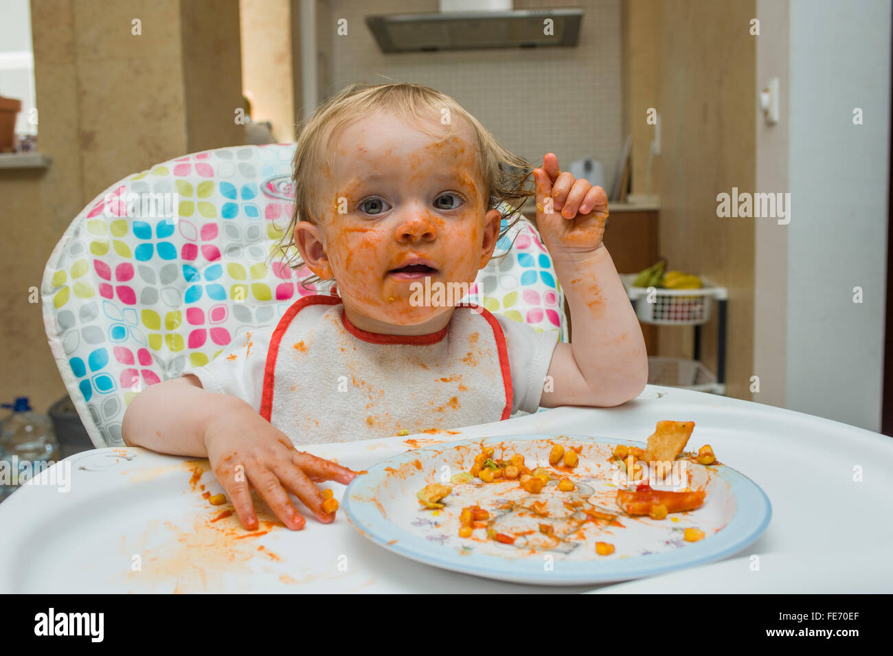 Una bambina (età 15 mesi) mangia un pasto a base di mais dolce e salsa di pomodoro in un modo disordinato e gioca con il suo cibo su una sedia alta Foto Stock