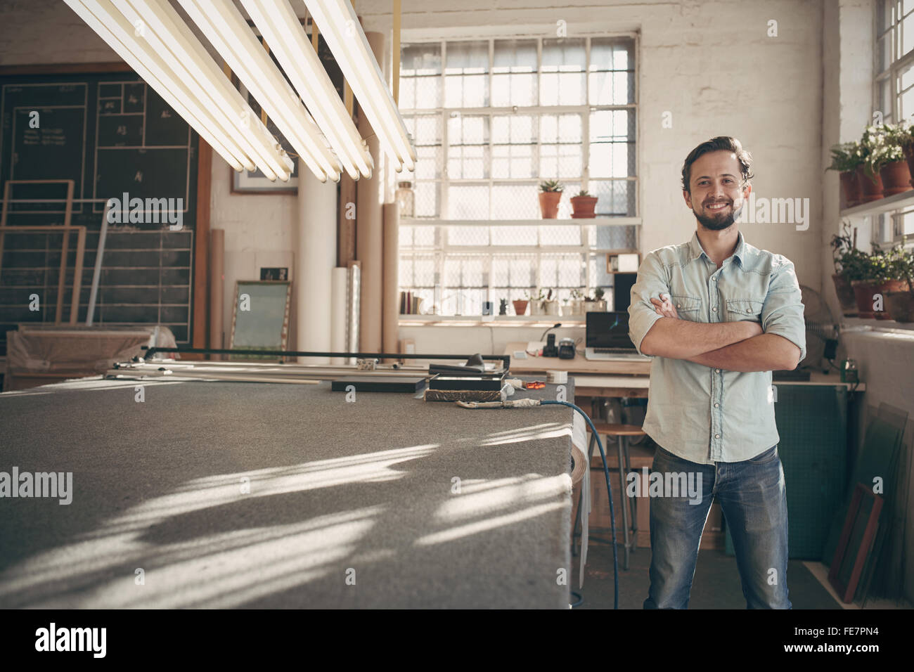 Ritratto di una buona ricerca designer maschio in piedi nella sua officina studio con le braccia piegate e sorridente con fiducia a camma Foto Stock