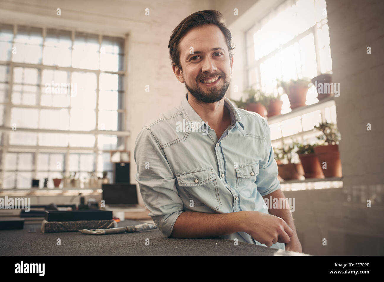 Ritratto di una giovane designer maschio appoggiano sul suo banco e dando la telecamera una rilassata e cordiale sorriso Foto Stock