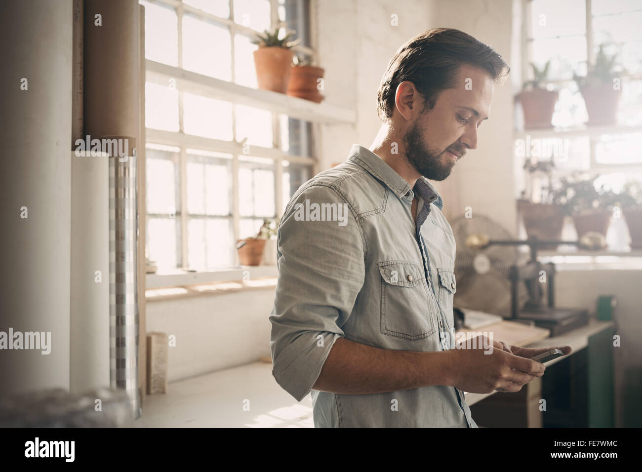 Bello imprenditore nel casual guardando al suo telefono mentre è in piedi la dolce luce della sua officina studio Foto Stock