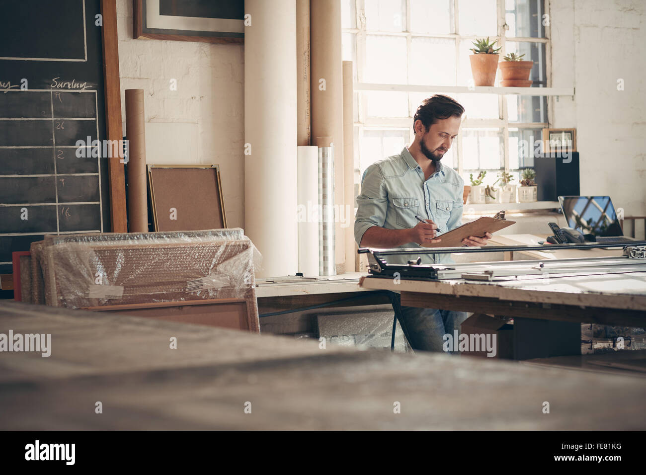 Maschio di imprenditore artigiano controllare gli ordini e le figure su un clipboard mentre in piedi casualmente nel suo studio di officina Foto Stock