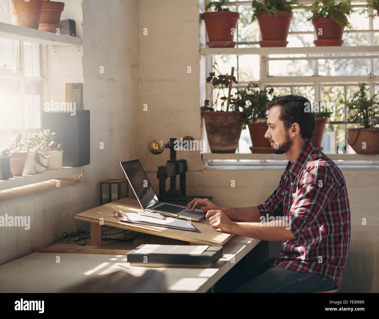 I giovani designer la digitazione sul suo computer portatile mentre è seduto in uno spazio creativo che egli utilizza come il suo ufficio nel suo studio Foto Stock