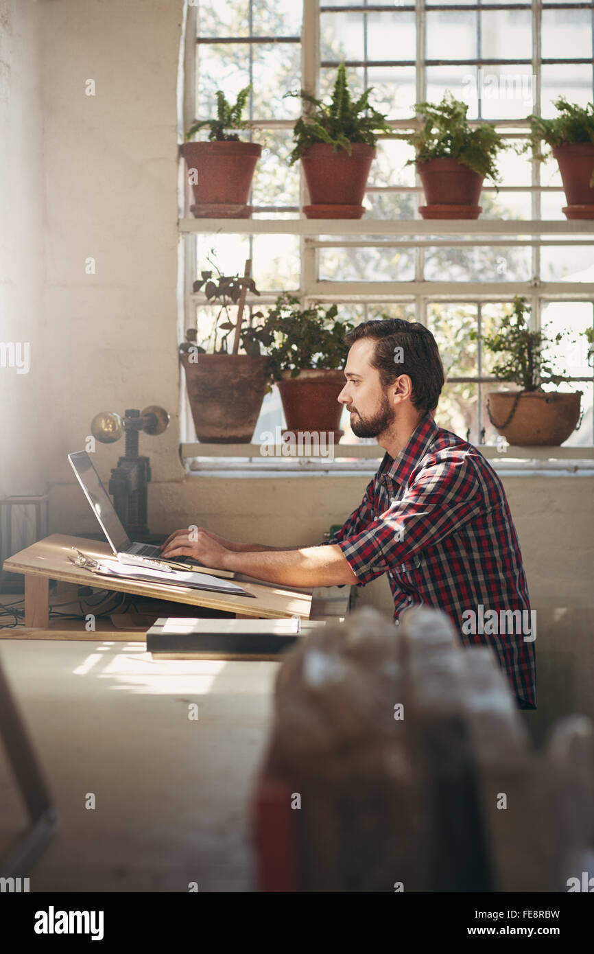 Giovane maschio imprenditore lavora con concentrazione sul suo computer portatile mentre è seduto in ufficio spazio del suo studio di design Foto Stock
