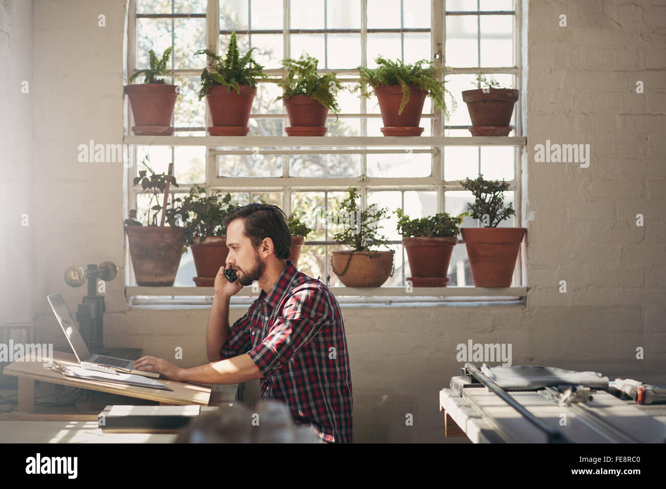 I giovani designer imprenditore guardando al suo computer portatile durante la conversazione sul suo telefono cellulare nel suo studio dello spazio di lavoro Foto Stock