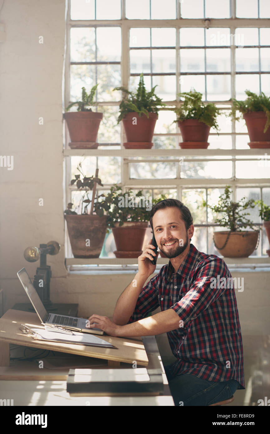 Imprenditore sorridente seduto alla scrivania del suo ufficio di studio e ricerca piacevolmente a la telecamera mentre ascolto la sua cella p Foto Stock