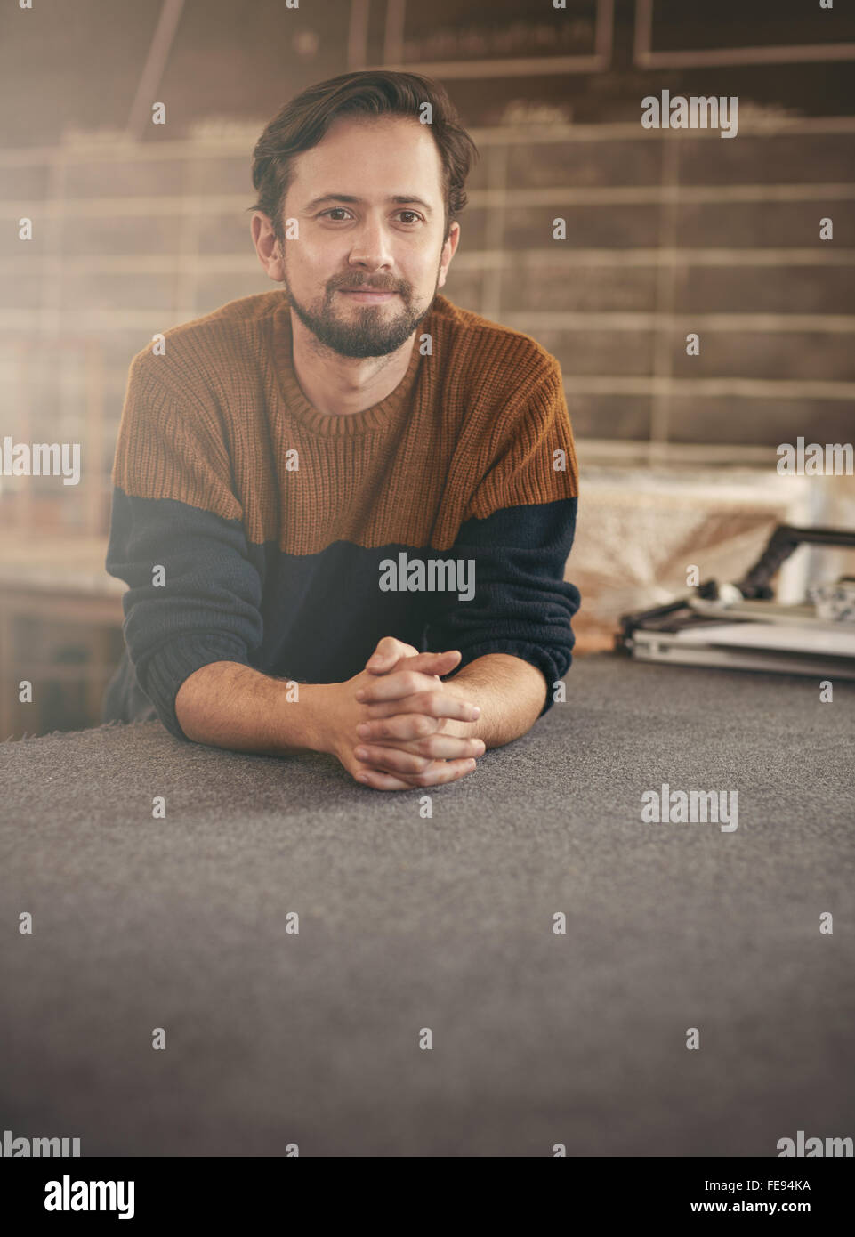 Casualmente vestito giovane uomo appoggiato su di una superficie di lavoro nel suo studio e guardando lontano in un positivo ancora pensieroso modo Foto Stock