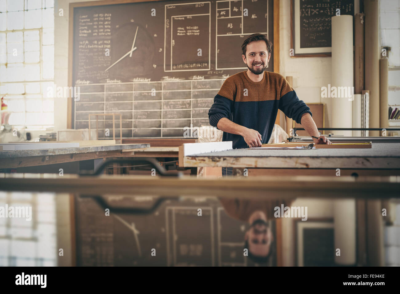 Artigiano guardando verso l'alto dal suo lavoro e sorridere mentre in piedi nel suo studio occupato con un progetto di falegnameria Foto Stock