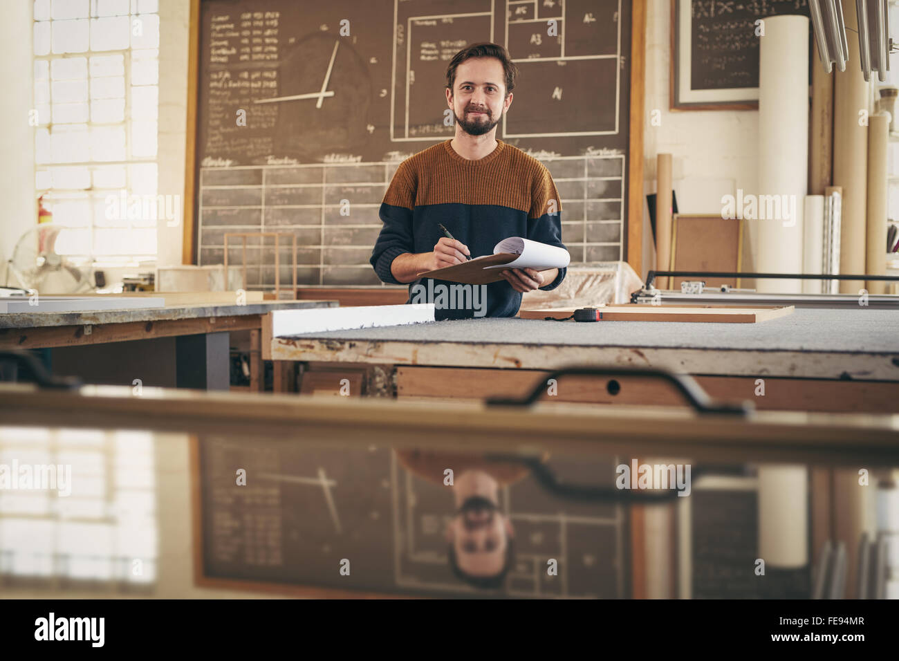Ritratto di un professionista artigiano sorridente alla videocamera mentre si lavora nel suo studio e utilizzando un blocco appunti e una penna per controllare st Foto Stock
