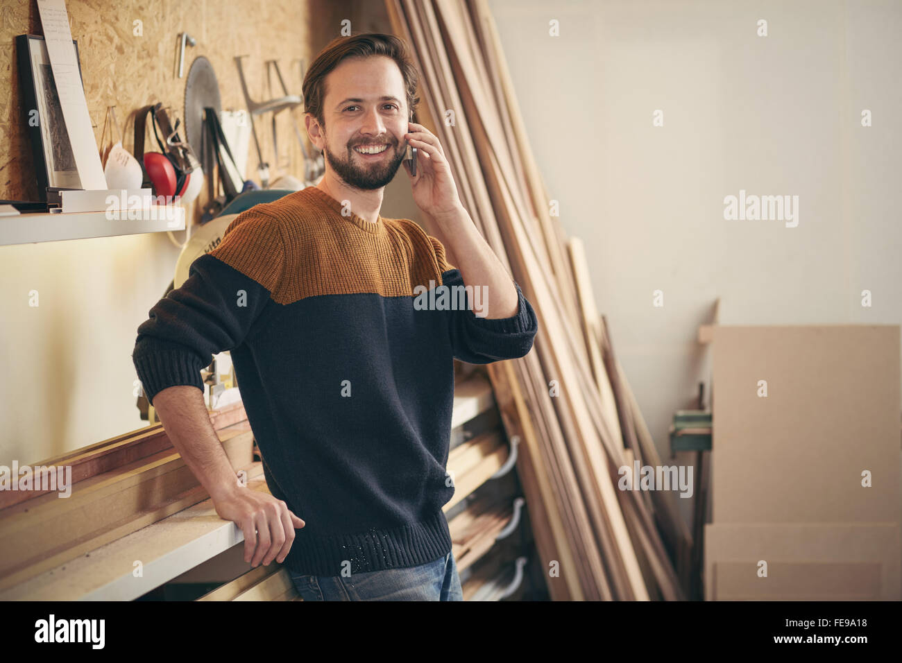 Ritratto di un giovane uomo sorridente mentre si parla sul suo telefono cellulare e in piedi casualmente nella sua officina per la lavorazione del legno Foto Stock