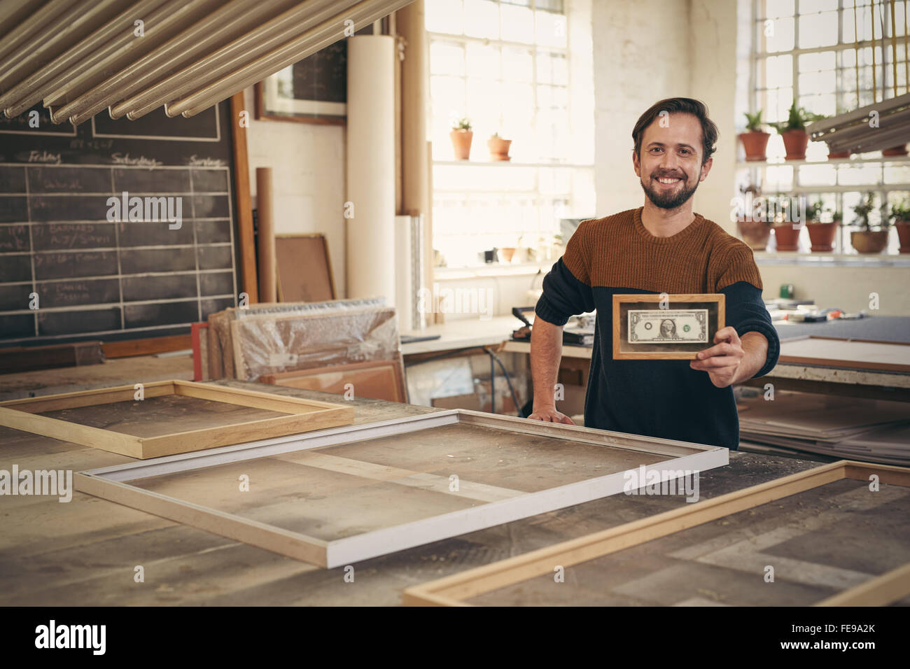 Il giovane imprenditore sorridente con orgoglio nel suo laboratorio mentre si tiene su una cornice per una nota banca Foto Stock