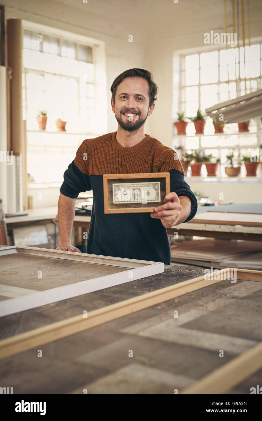 Giovane artigiano tenendo in mano una banca nota che è stata inquadrata mentre si erge con orgoglio nel suo laboratorio Foto Stock