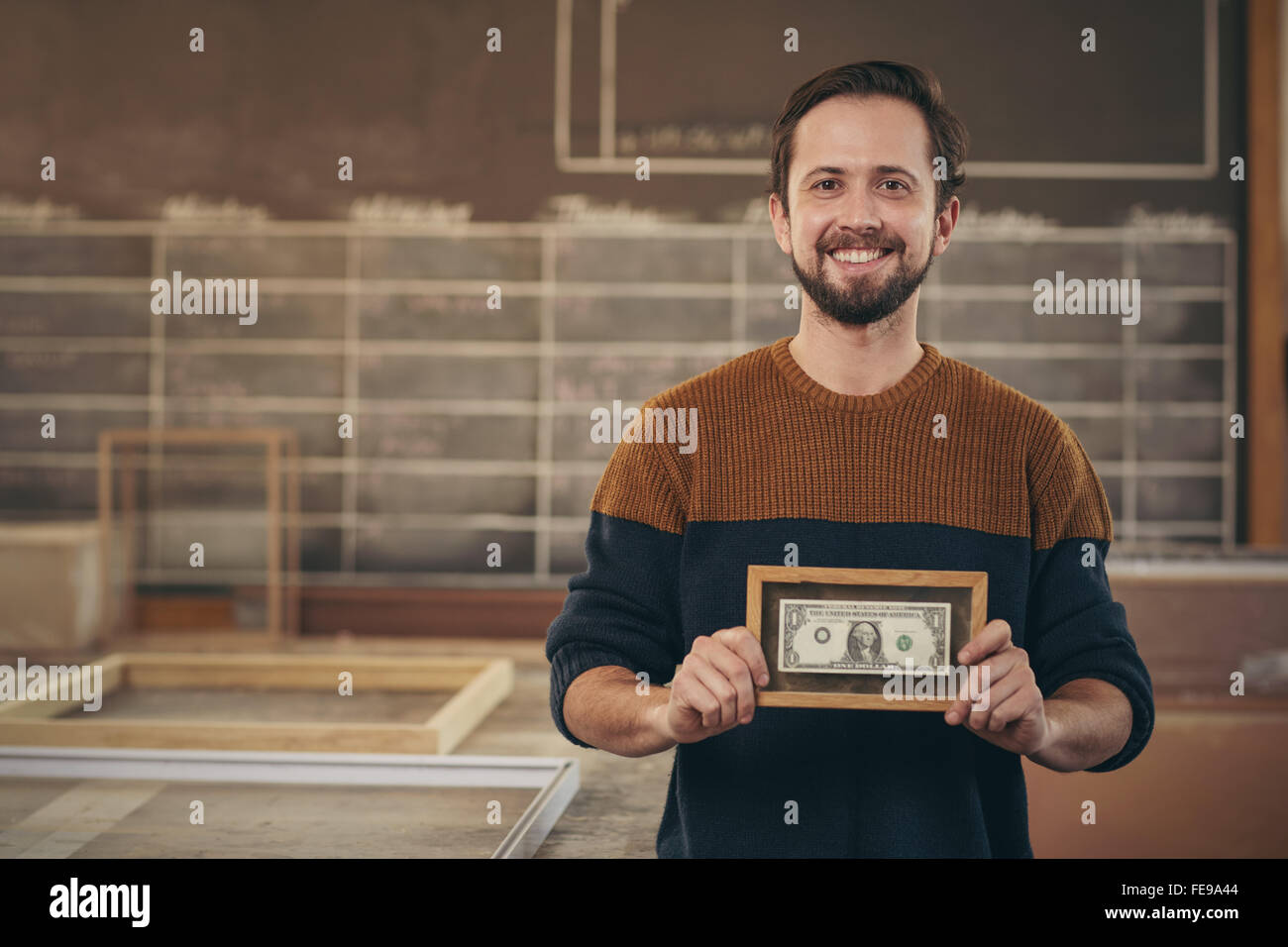 Sorridente imprenditore artigiano erge orgogliosamente nel suo laboratorio e la visualizzazione di una banca nota che è stata accuratamente incorniciato Foto Stock