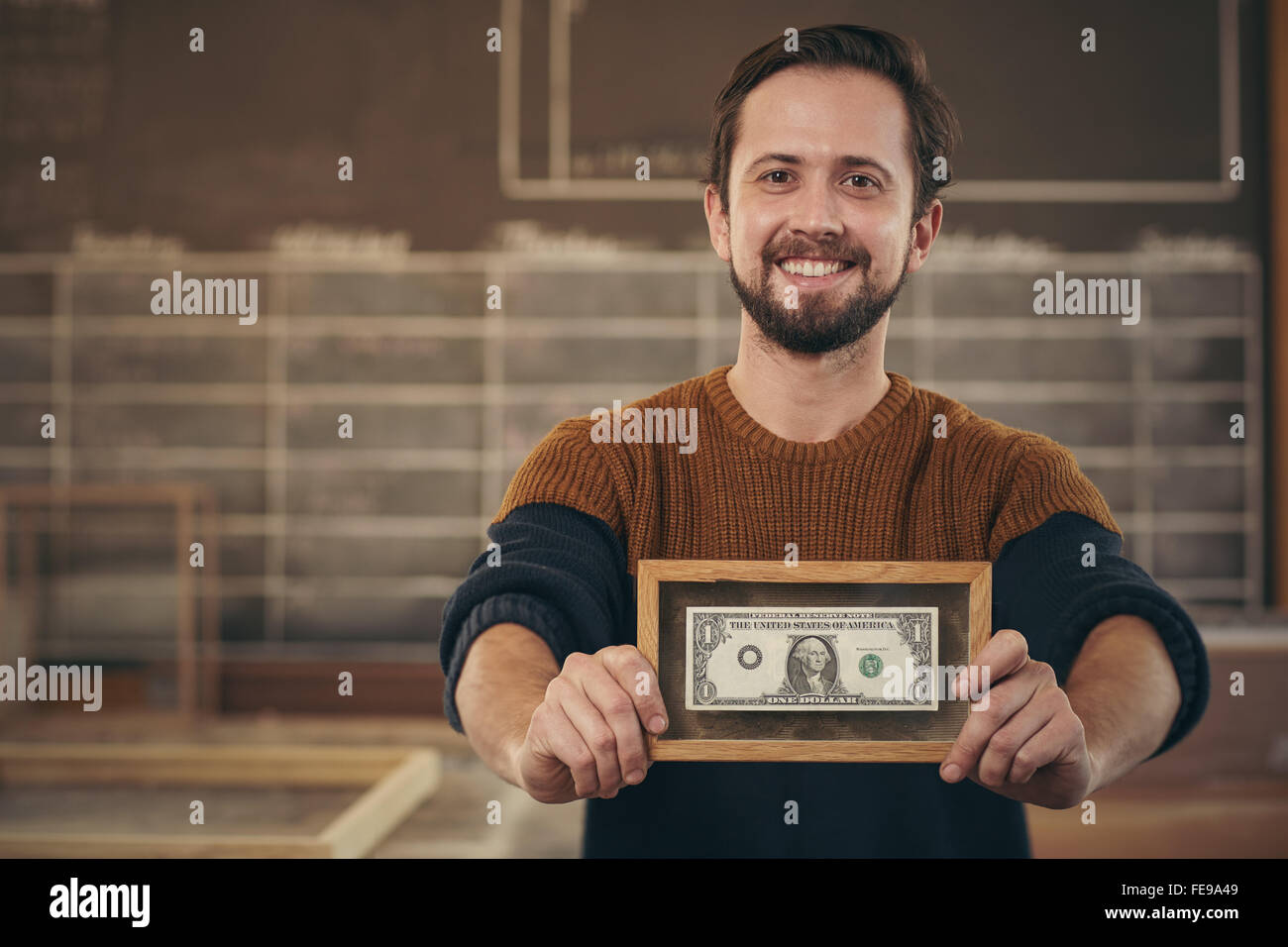 Imprenditore sorridente in piedi con orgoglio nel suo laboratorio e vi mostra una banca nota che è stata elaborata Foto Stock