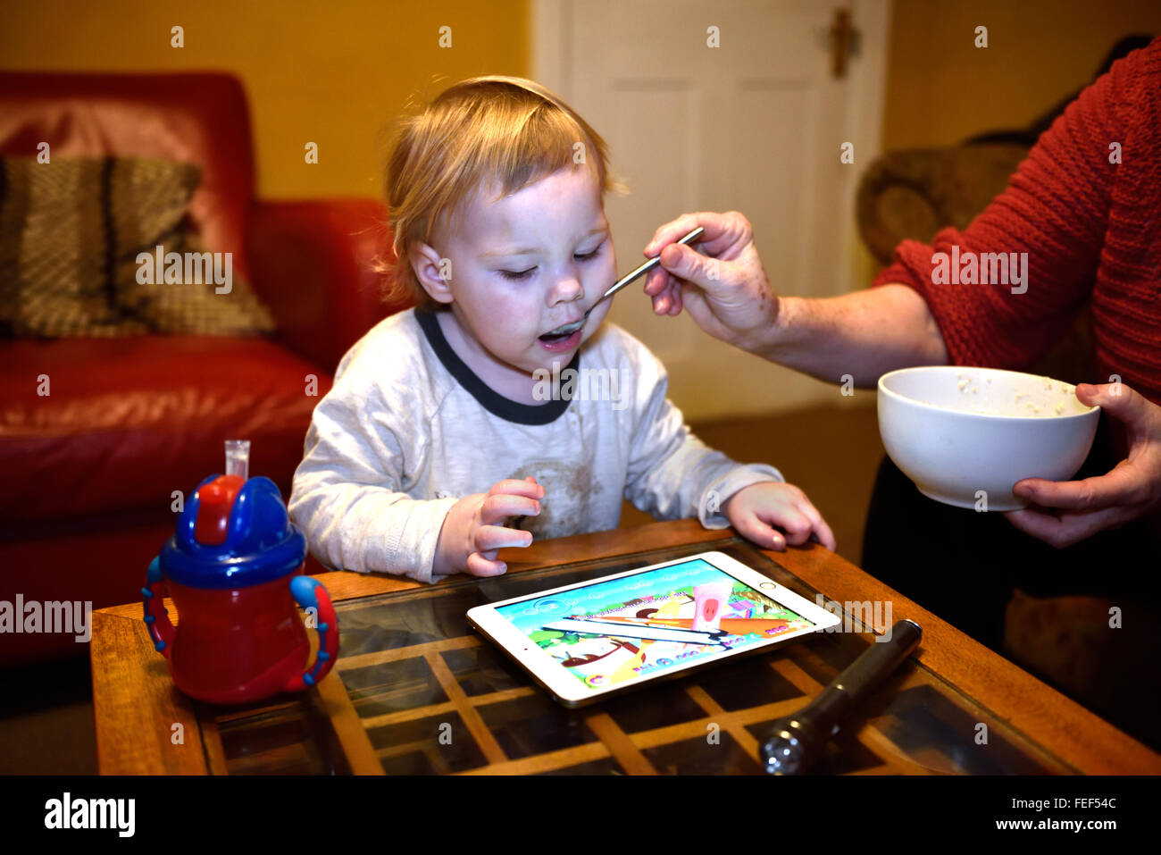 Un diciotto mesi essendo toddler Spoon Fed durante la riproduzione di un gioco sul suo ipad nella sua casa. Foto Stock