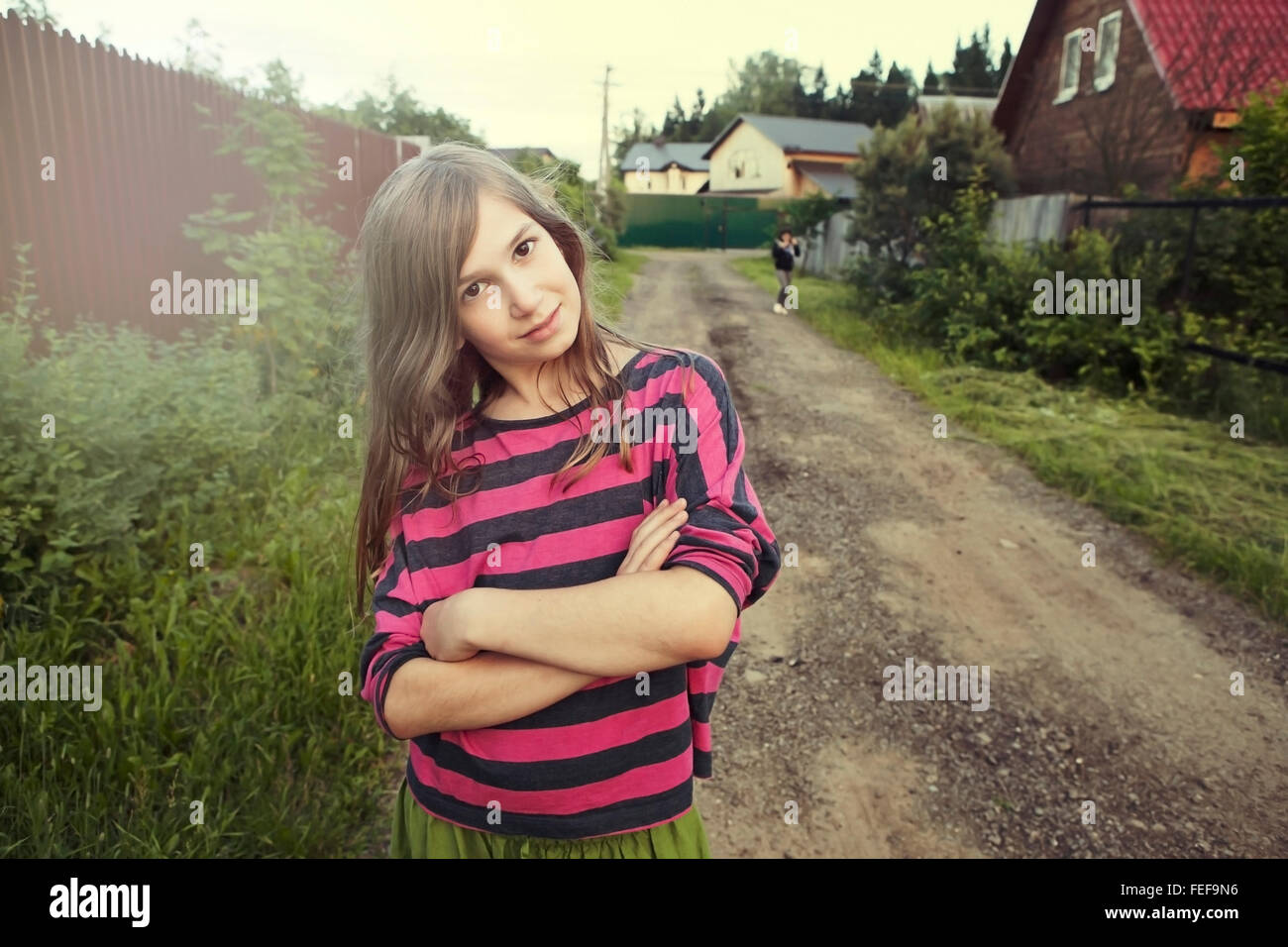 Sorridente ragazza adolescente in un rosso striped shirt permanente sulla strada tra le case, le braccia incrociate sul petto. Foto Stock