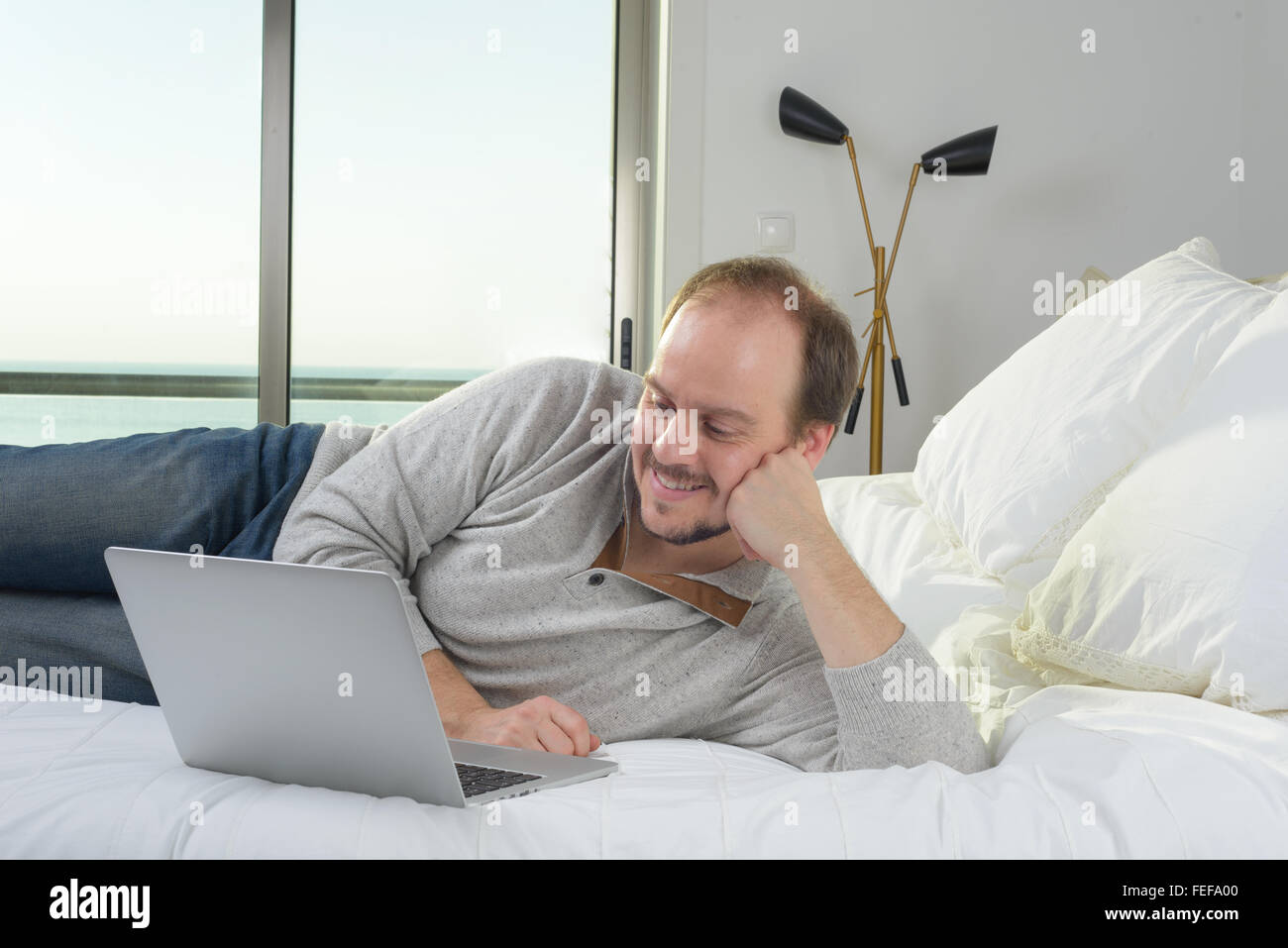 Uomo disteso sul letto sorridente lavorando sul computer portatile Foto Stock