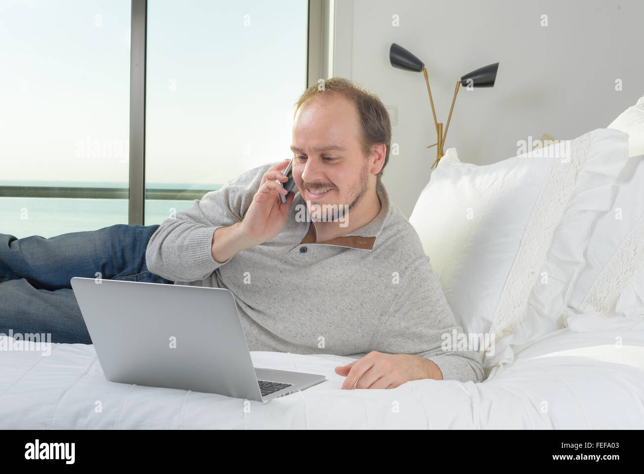 Uomo disteso sul letto sorridente lavorando sul computer portatile parlando al cellulare mobile Foto Stock