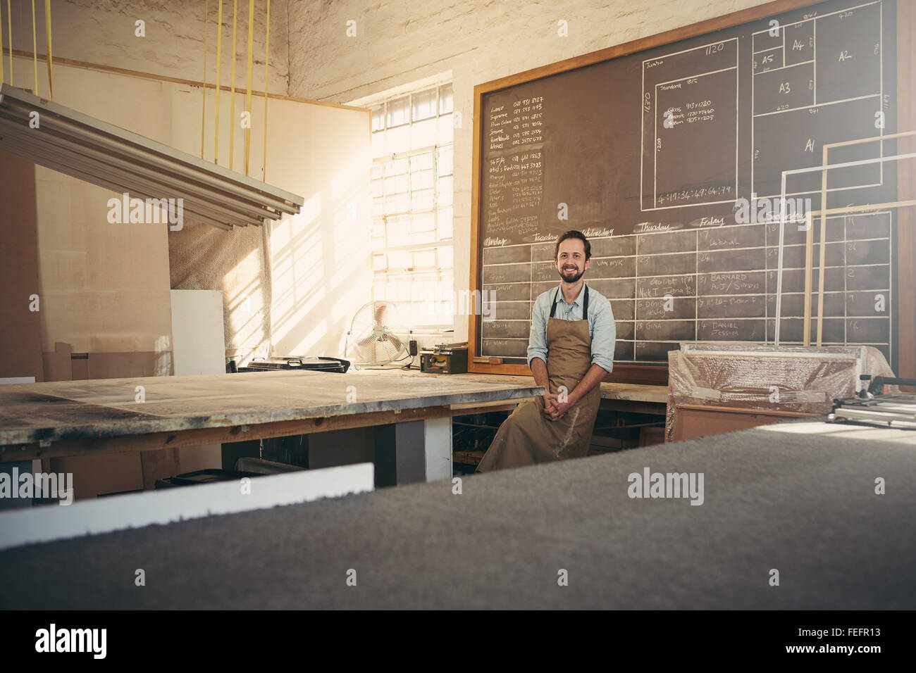 Ritratto di bello falegname artigiano sorridente alla telecamera mentre appoggiata contro il suo banco di lavoro nella sua officina Foto Stock